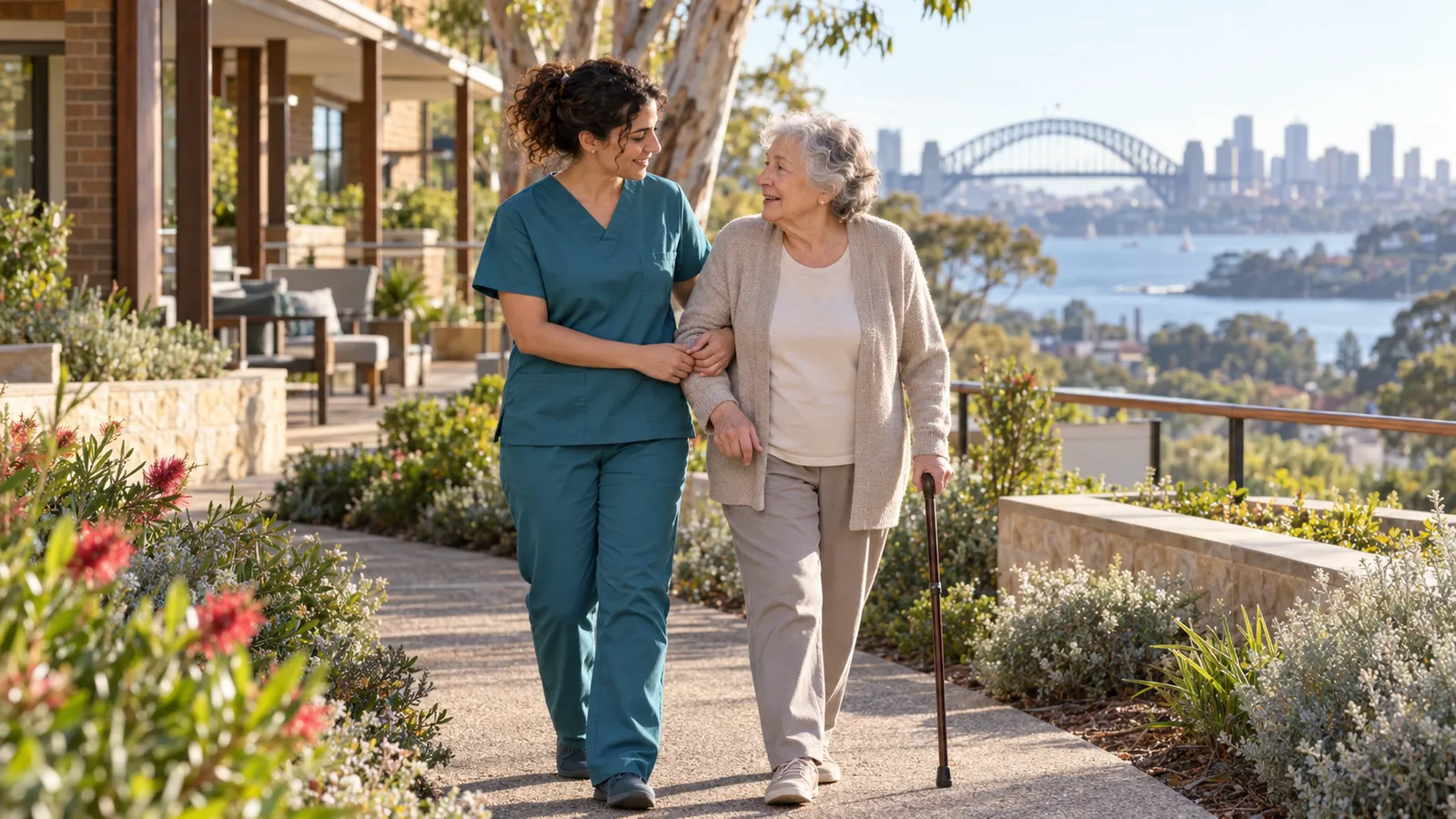 Una giovane operatrice accompagna un’anziana in un giardino soleggiato, con il porto di Sydney sullo sfondo.