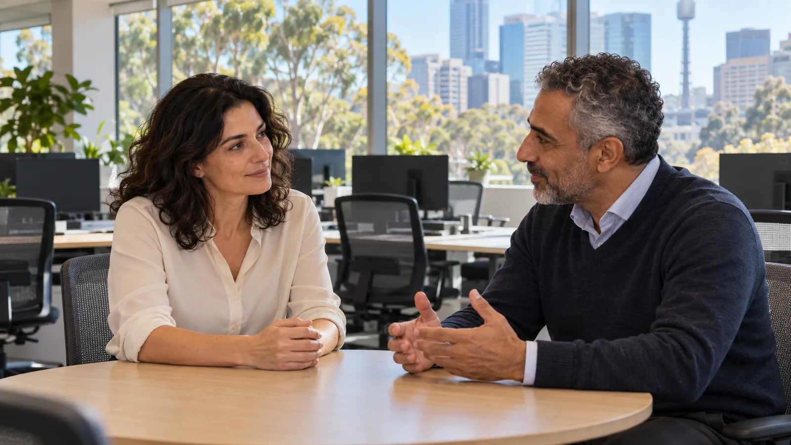 Due professionisti italiani discutono con calma in un ufficio australiano luminoso con postazioni vuote.